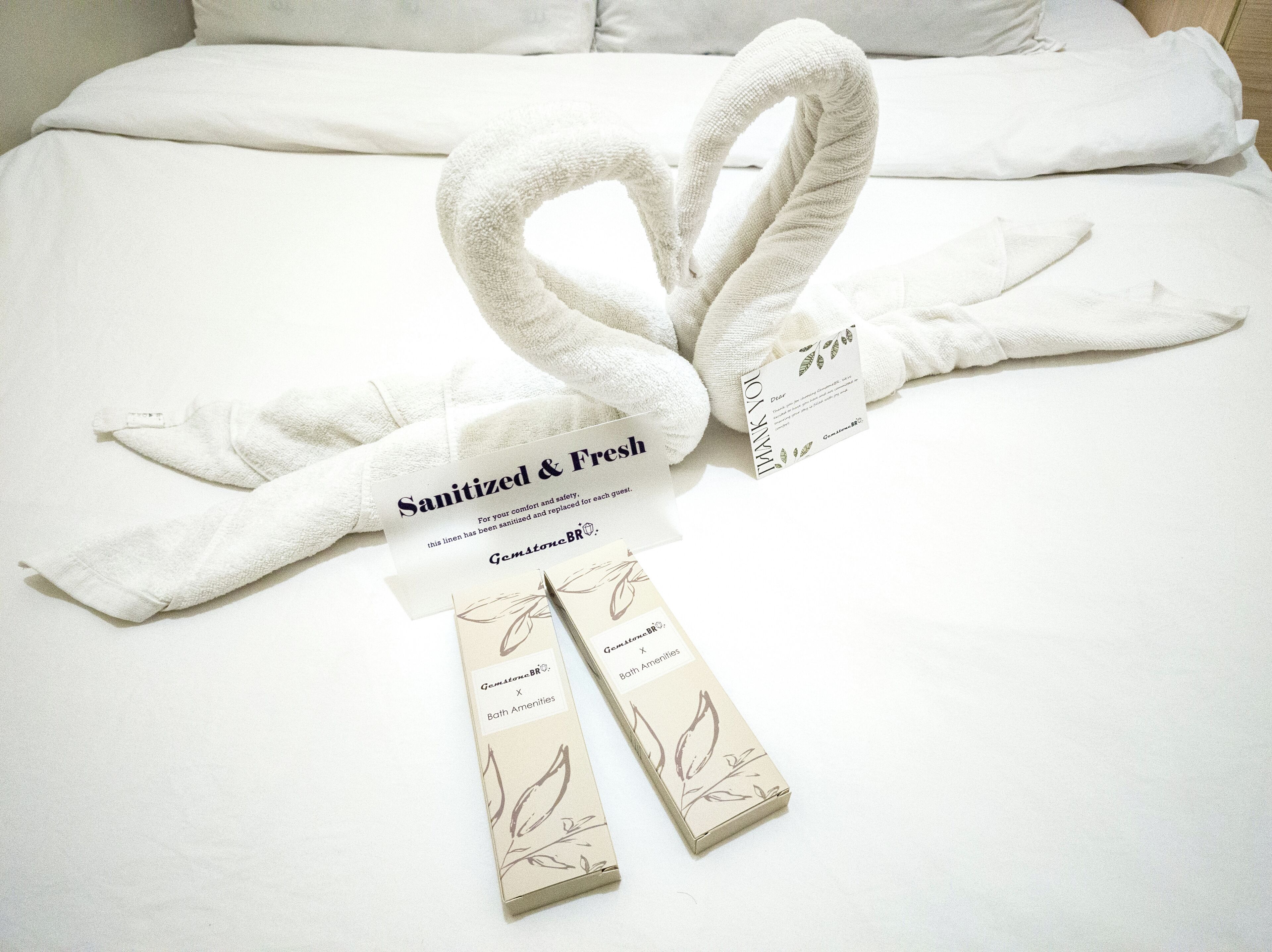 A neatly made bed with white linens, a heart-shaped towel, and a tag indicating the linens are sanitized and fresh.