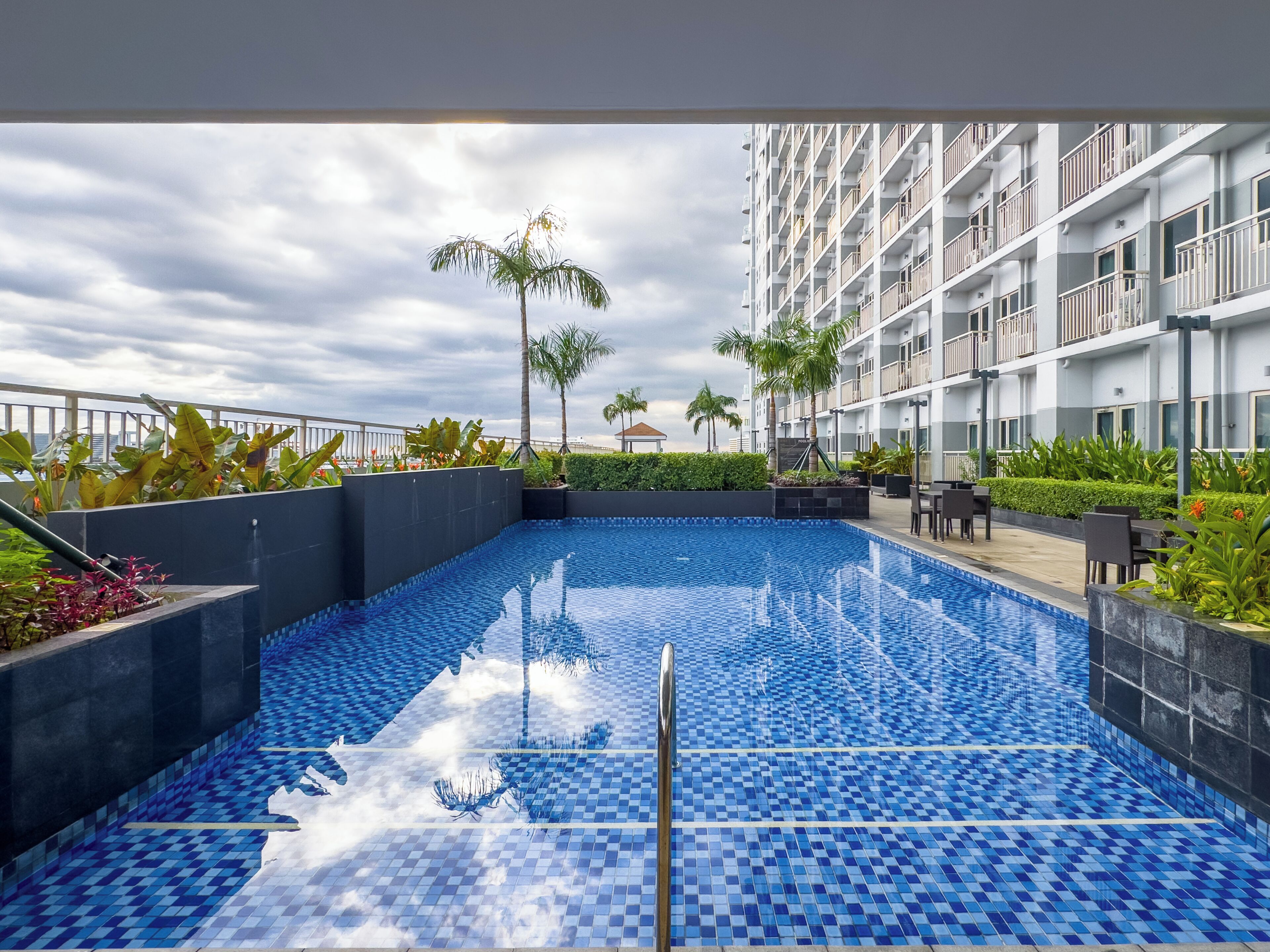 A swimming pool with blue tiles, surrounded by greenery and a multi-story building with balconies.