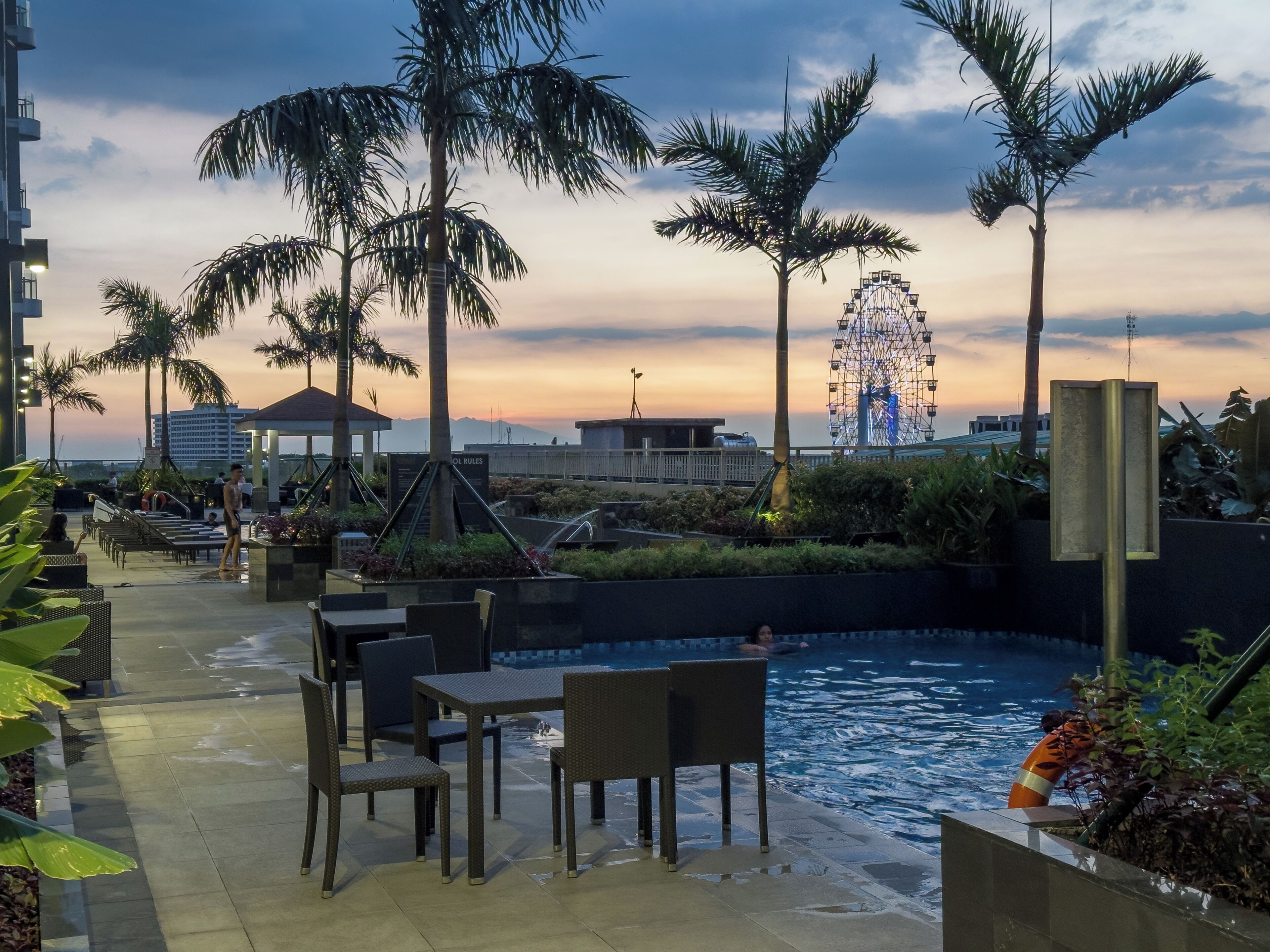 Een zwembad met stoelen en tafels, een reuzenrad op de achtergrond en palmbomen.
