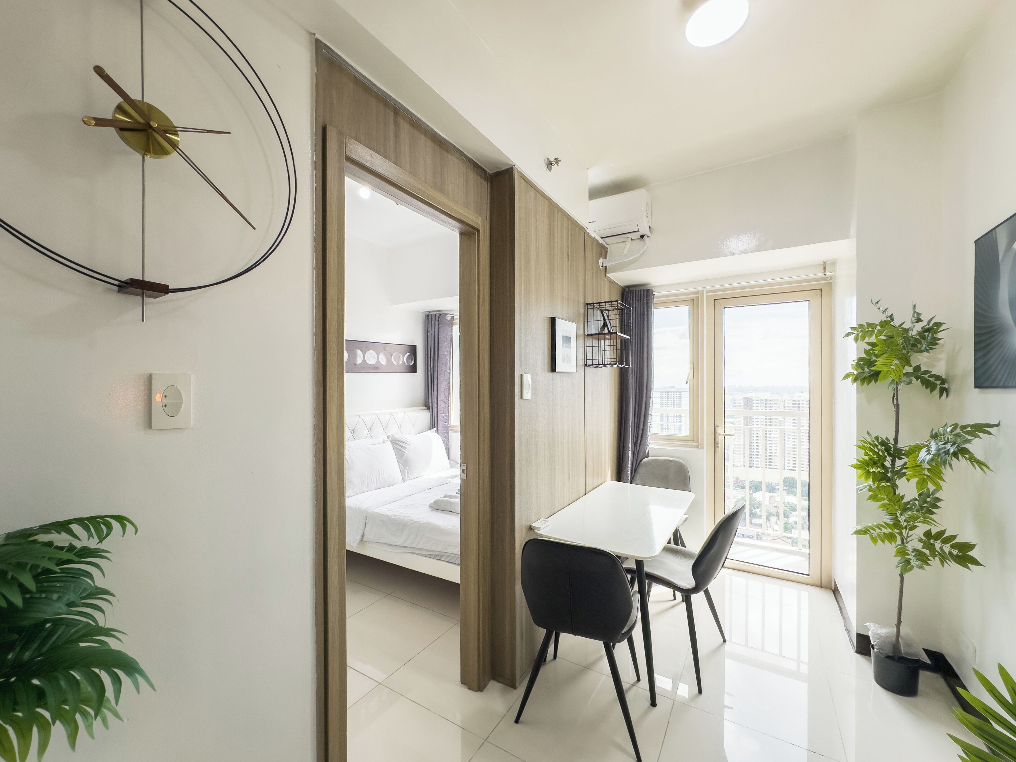 A modern hotel room with a bed, a dining area with a table and chairs, a clock on the wall, and a potted plant.