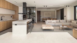 A modern kitchen with a central island, bar stools, and a dining area with a sofa and coffee table. - Sancy Luxury Villa 400m beach, Heated* pool & Gym (Chania)