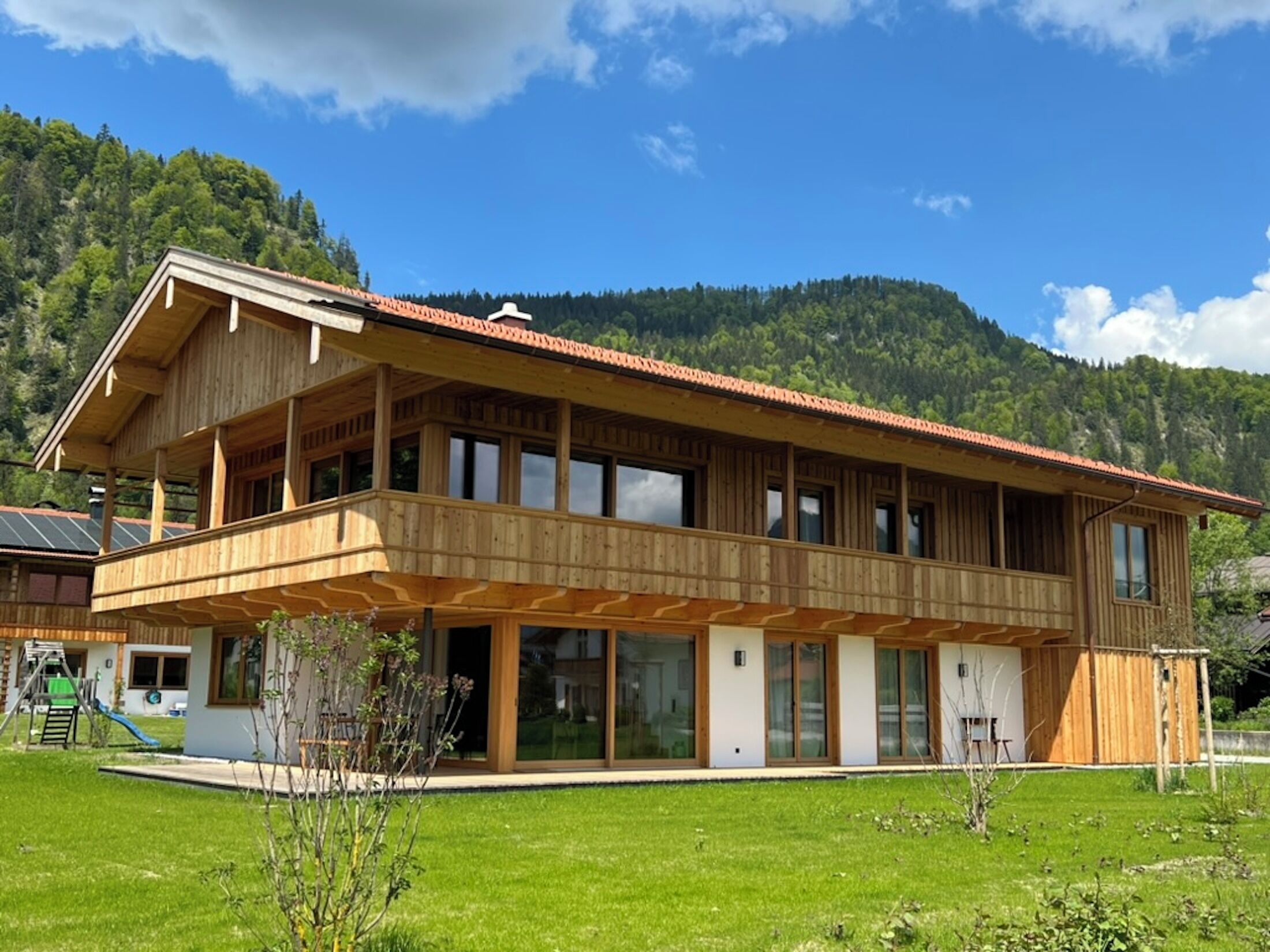 A two-story wooden house with a balcony, large windows, and a mountain backdrop.