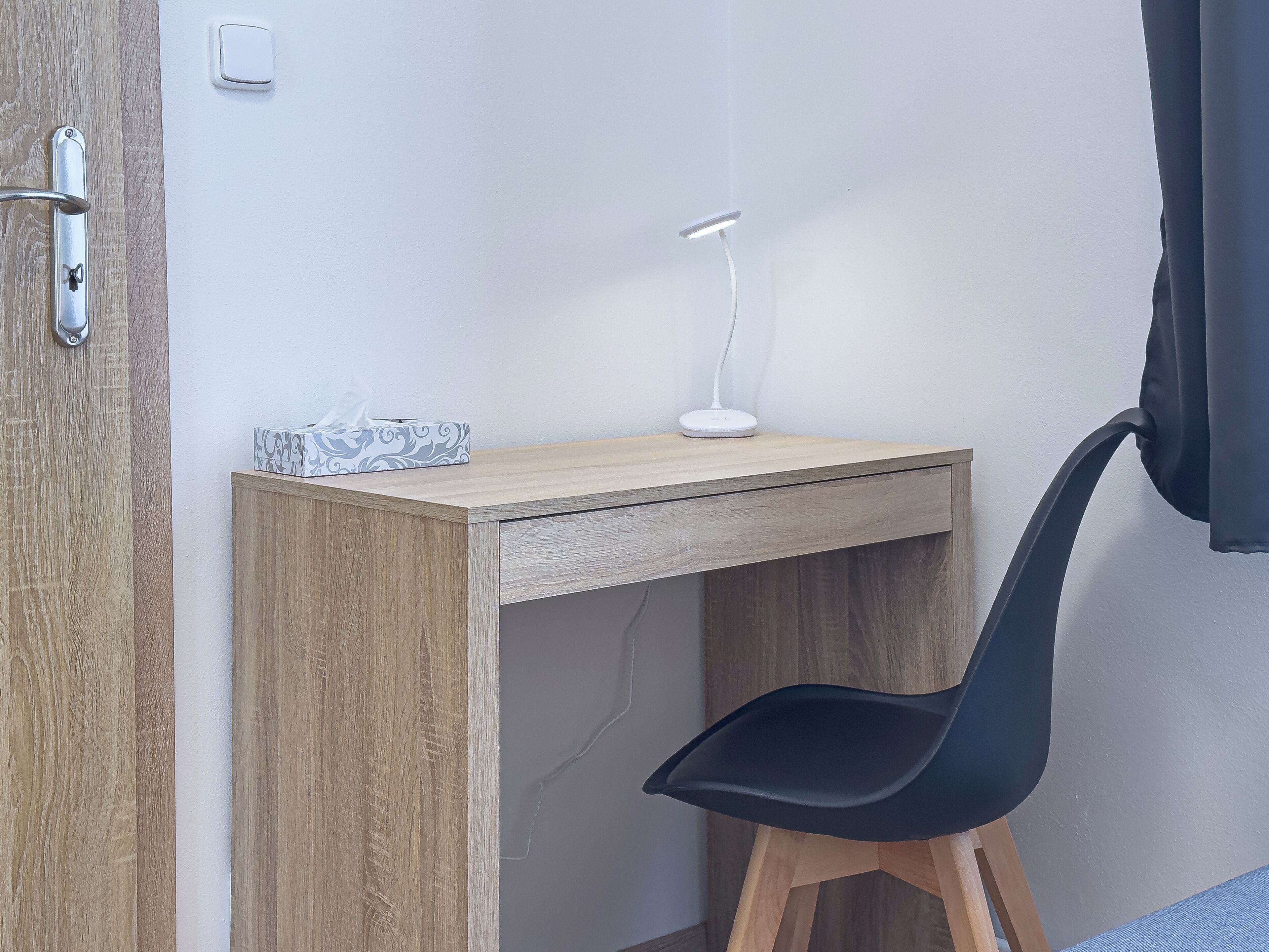 Een houten bureau met een witte bureaulamp, een zwarte stoel en een zakdoosje.
