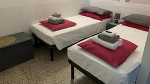 A hotel room with two single beds, each with a red pillow and a folded towel. There is a small bedside table with a lamp and a decorative item. The room has a textured white wall and a geometric wall art piece. - Hotel Splendor (Tortoli)