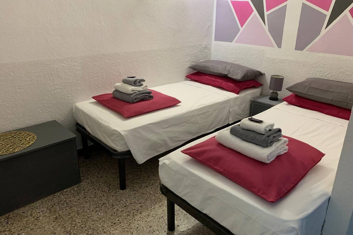 A hotel room with two single beds, each with a red pillow and a folded towel. There is a small bedside table with a lamp and a decorative item. The room has a textured white wall and a geometric wall art piece.