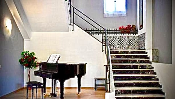 A grand piano in a spacious room with a wooden floor, a staircase with patterned railings, and a small table with a plant. - Hotel Splendor (Tortoli)
