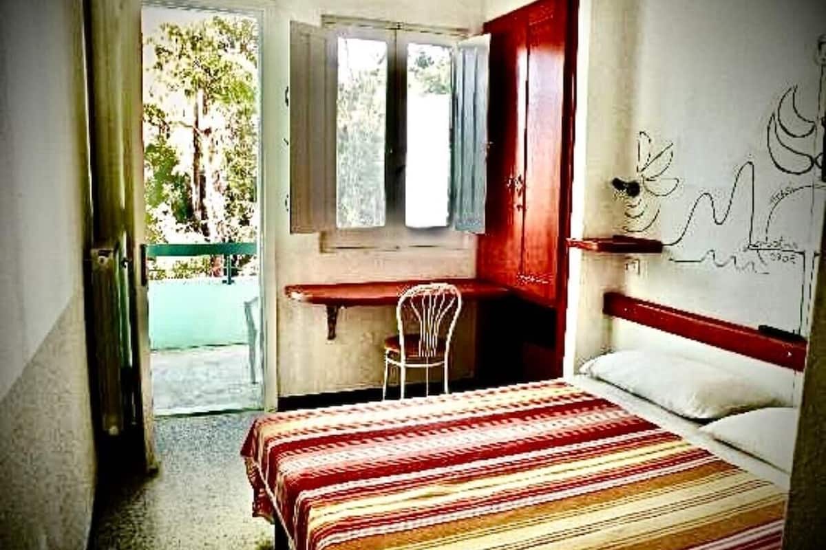A bedroom with a bed, a desk, and a chair. There is a window with a view of trees.