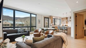 A spacious living area with a large window, a sofa, a coffee table, and a dining area with chairs and a table. - One Riverfront Condo 207 (Avon)