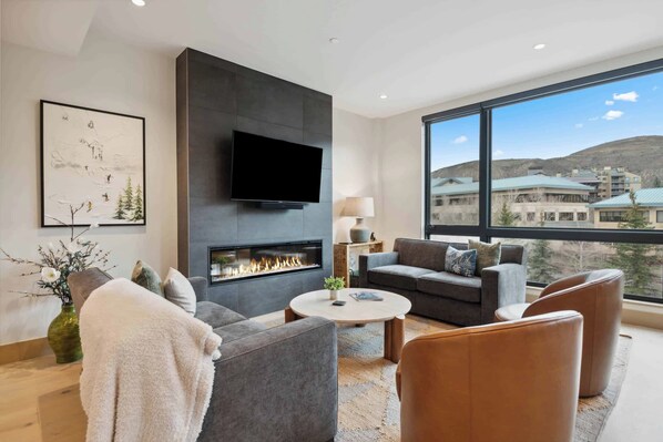 A modern living room with a fireplace, a flat-screen TV, and a large window offering a view of buildings and hills. - One Riverfront Condo 207 (Avon)