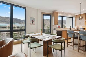 A modern kitchen with a large window, a dining area with green chairs, and a view of a mountainous landscape. - One Riverfront Condo 207 (Avon)