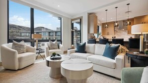 A modern living room with a large window, a sofa, a coffee table, and a kitchen area with pendant lights. - One Riverfront Condo 306 (Avon)