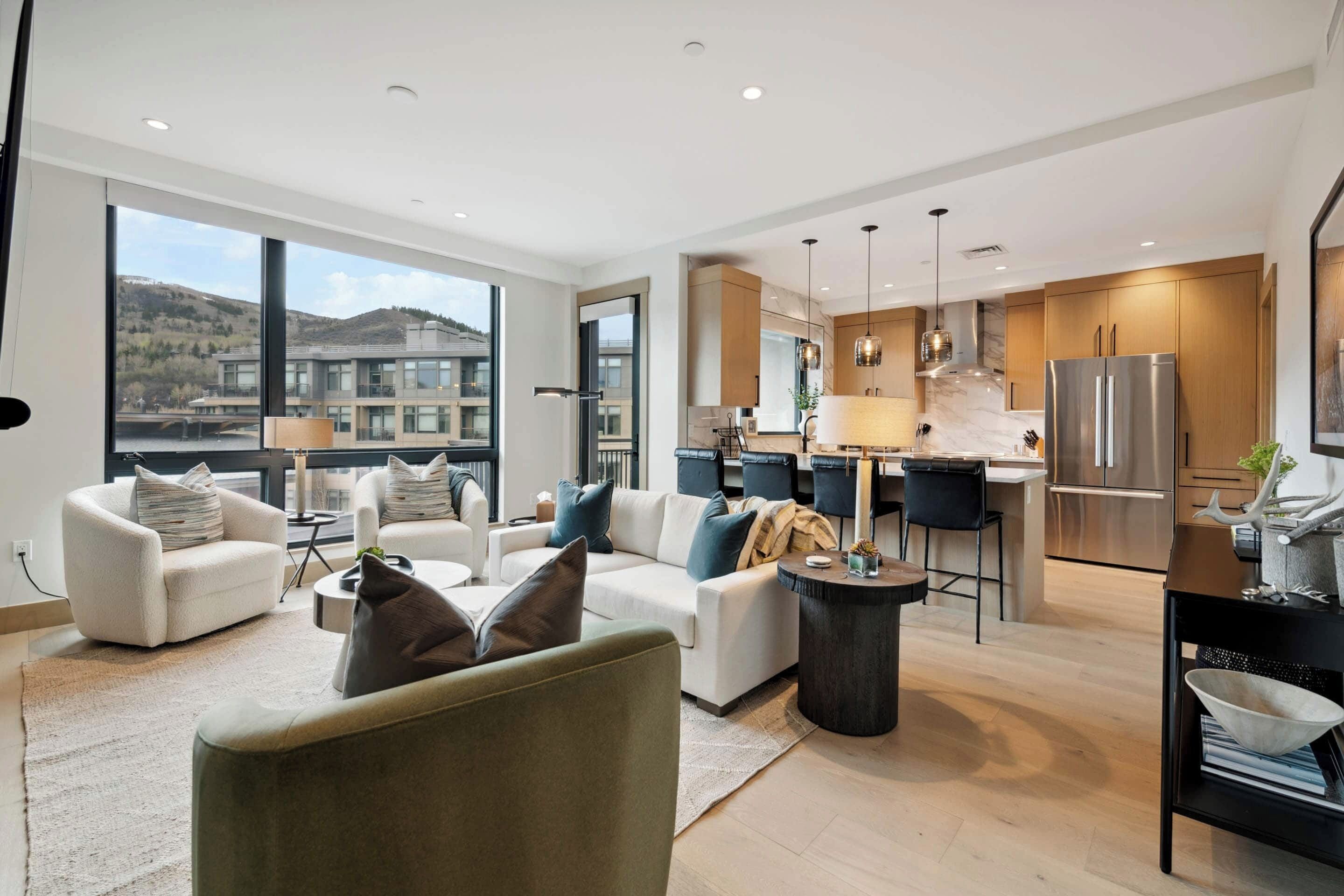 Una sala de estar moderna con sofá, sillones y una mesa de centro. Cuenta con una zona de cocina con refrigerador y una mesa de comedor. La habitación tiene ventanas grandes y vistas al exterior.
