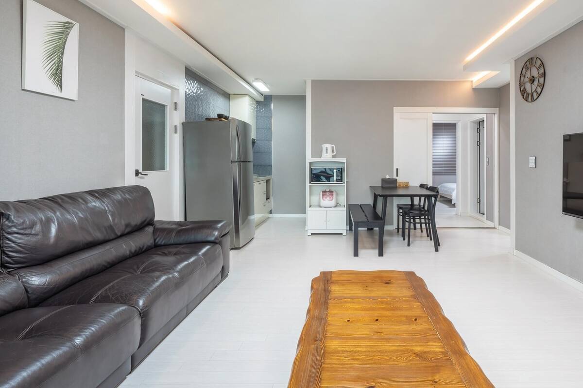 Una sala moderna con un sofá de cuero, una mesa de centro de madera, un comedor con mesa y sillas, una cocina con refrigerador y microondas, y un televisor montado en la pared.