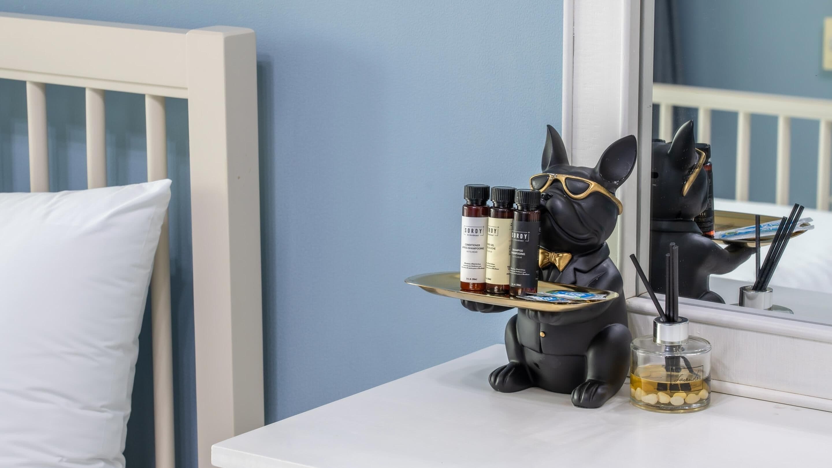 Una figurita de gato negro sobre una bandeja que sostiene dos botellas de SORDY para el cuerpo y una botella de aceite corporal SORDY, con un vaso de aceite y un par de palillos negros a su lado, sobre una mesita de noche blanca.
