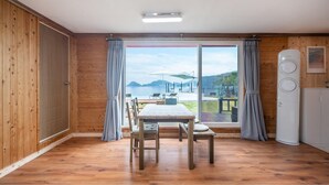 Una habitación con piso de madera, una mesa con sillas, un ventanal con cortinas y un electrodoméstico blanco a la derecha.