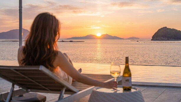 Una persona descansando en una silla de salón con una mesa, una botella y una copa de vino, con vista a un mar tranquilo y montañas al fondo.