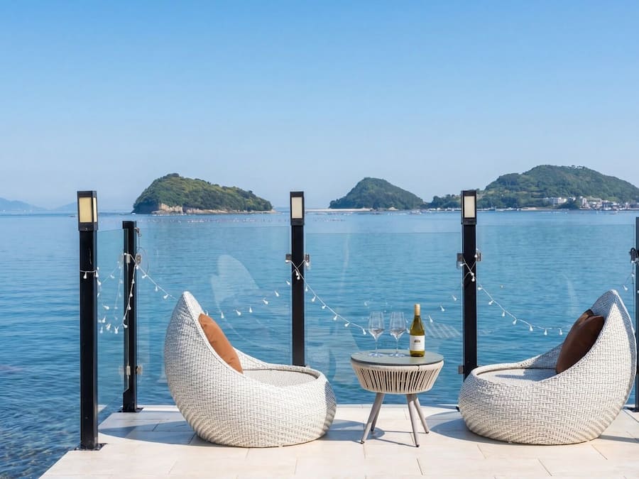 A seaside deck with two wicker chairs, a small round table with a bottle, and a view of the sea and distant islands.