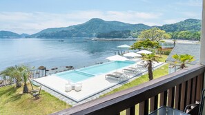 Una piscina con sillas de descanso y sombrillas, rodeada por una terraza de madera y palmeras, con vista a un lago tranquilo y montañas al fondo.