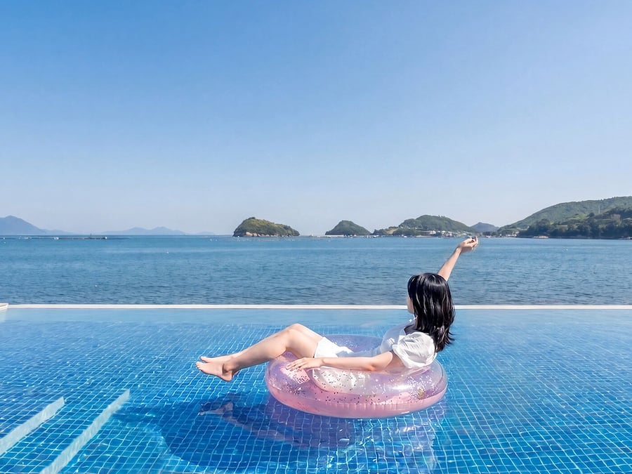 A person on a pink float in a clear blue pool with a view of the sea and distant islands.
