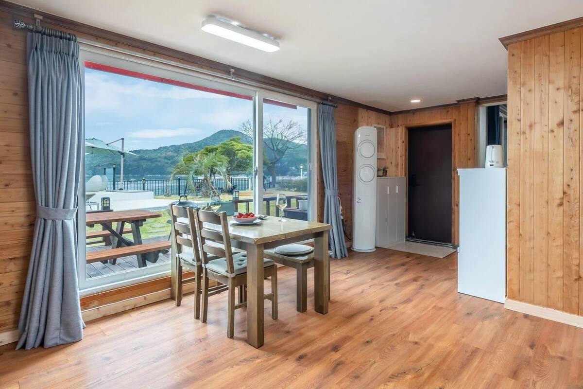 Una habitación con piso de madera, una mesa de comedor con sillas, un ventanal con cortinas y vistas a un parque y montañas.