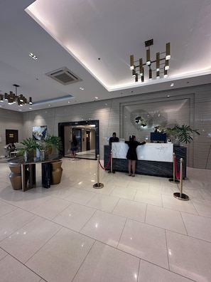 Eine moderne Hotelhalle mit Empfangstresen, Zimmerpflanzen, einer Marmor-Apotheke und einem großen, abstrakten Kunstwerk an der Wand.
