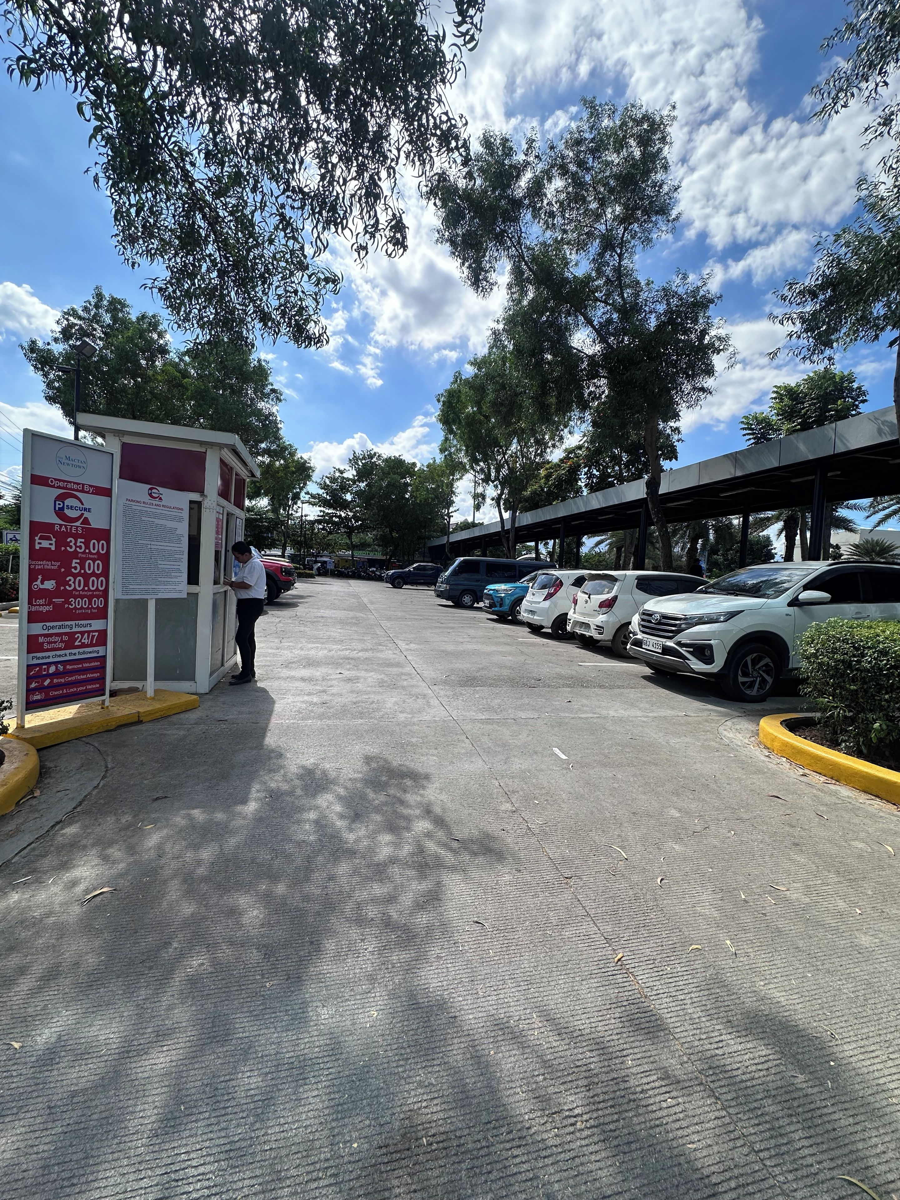 Un estacionamiento con una caseta de seguridad, coches aparcados y árboles.