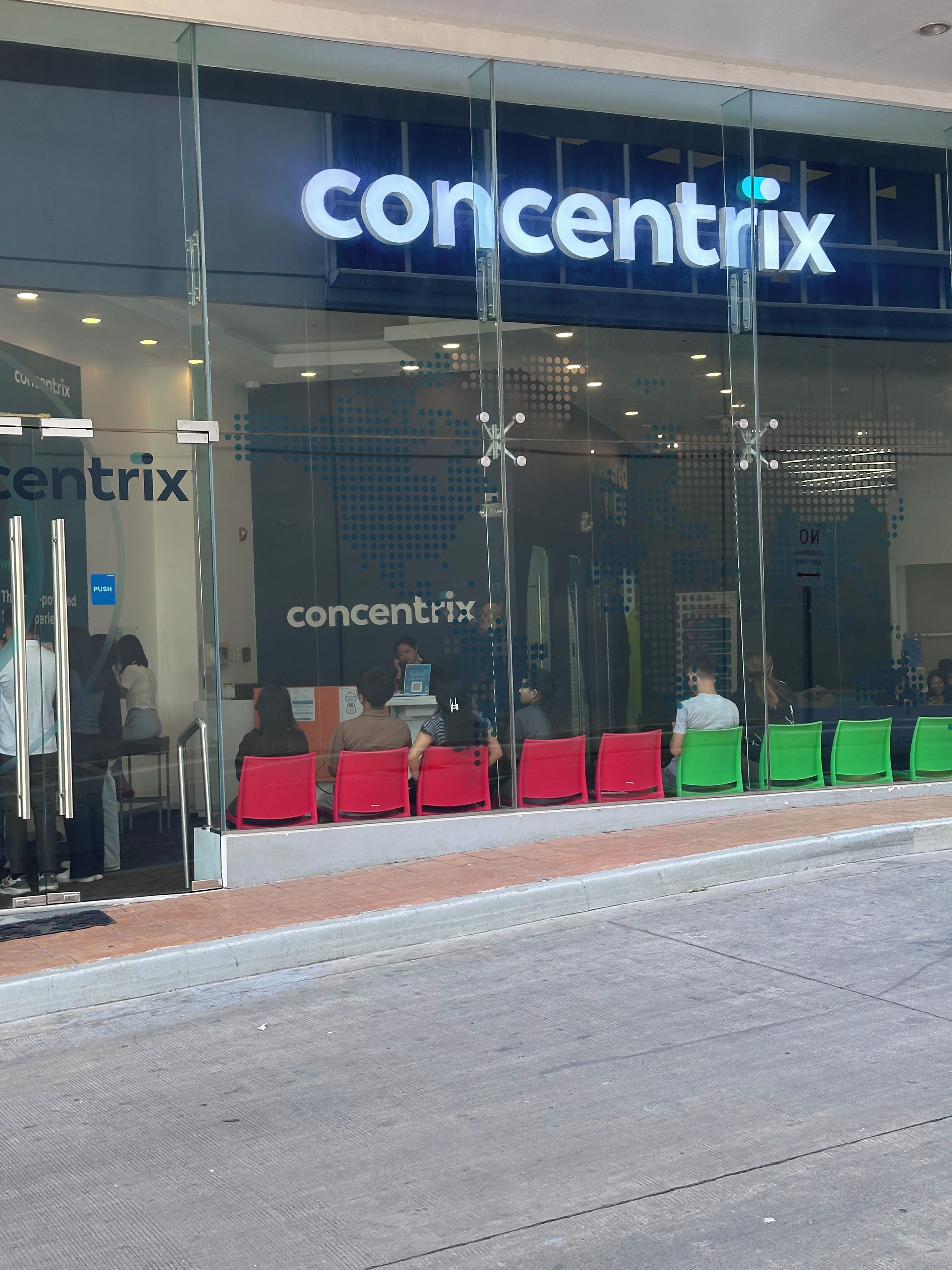 Un magasin Concentrix avec une façade en verre, des chaises rouges et vertes à l’extérieur et une entrée bien visible.