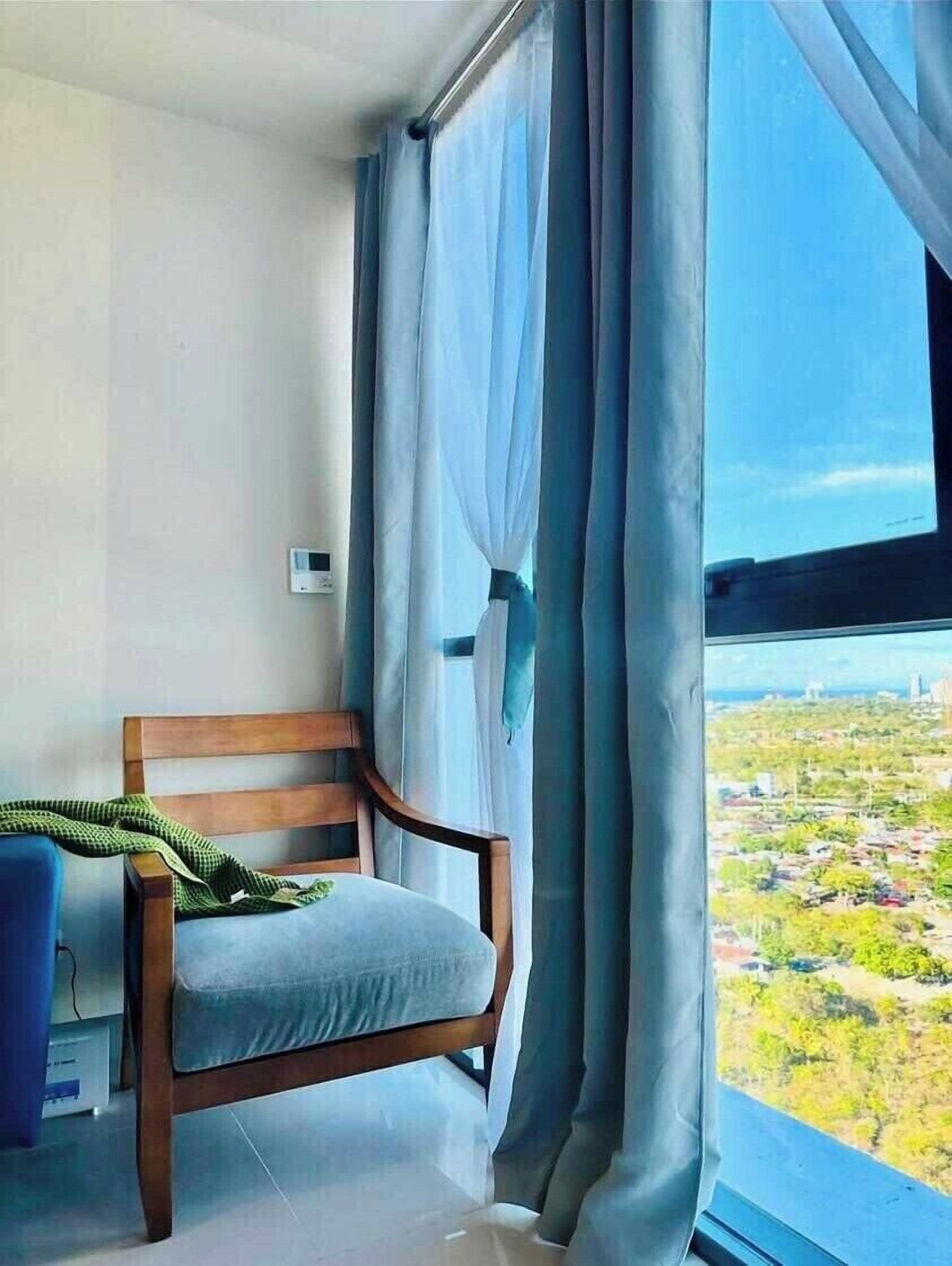 A room with a wooden chair, a view of a cityscape through large windows, and a curtain partially drawn to the side.