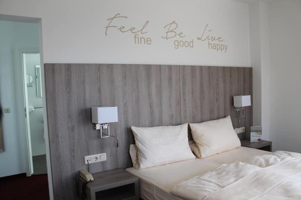 Ein Schlafzimmer mit hölzernem Kopfteil, zwei Nachttischen, einem Bett mit weißer Bettwäsche und einem motivierenden Zitat an der Wand.