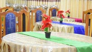 Un comedor con mesas redondas cubiertas de plástico, sillas de madera con tapas de vidrio azul y flores rojas sobre las mesas.