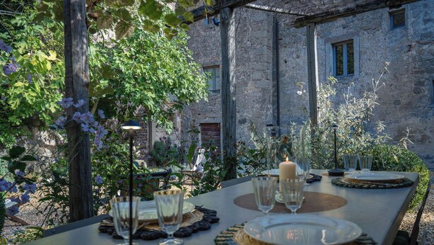 Un área de comedor al aire libre con una pared de piedra rústica de fondo, una mesa dispuesta con platos, vasos y una vela, rodeada de follaje y una pérgola.