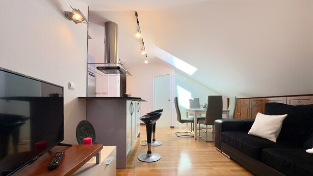 A modern living room with a flat-screen TV, a black sofa, a dining table, and a kitchen area with a stove and cabinets.
