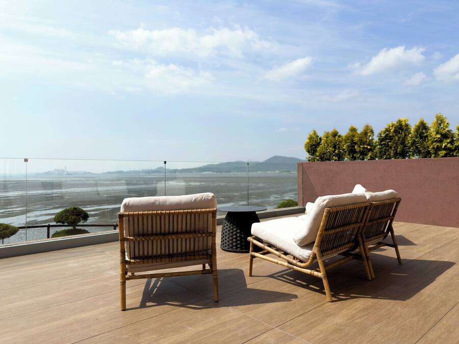 A balcony with two wicker lounge chairs and a small table, overlooking a body of water and distant cityscape.