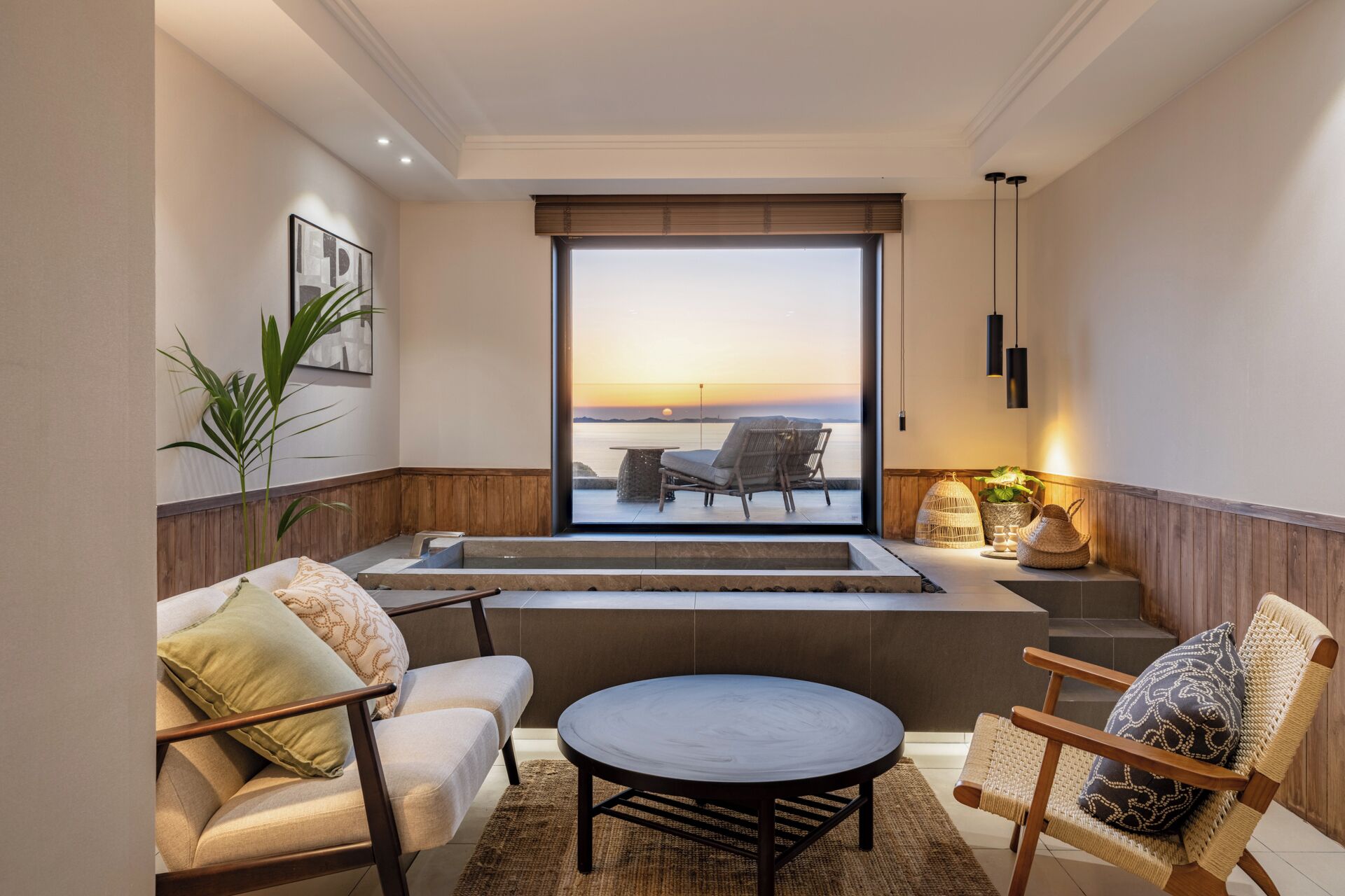 A modern living room with a large window overlooking a body of water at sunset, furnished with a sofa, chairs, and a coffee table.