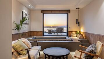 A modern living room with a large window overlooking a body of water at sunset, furnished with a sofa, chairs, and a coffee table.