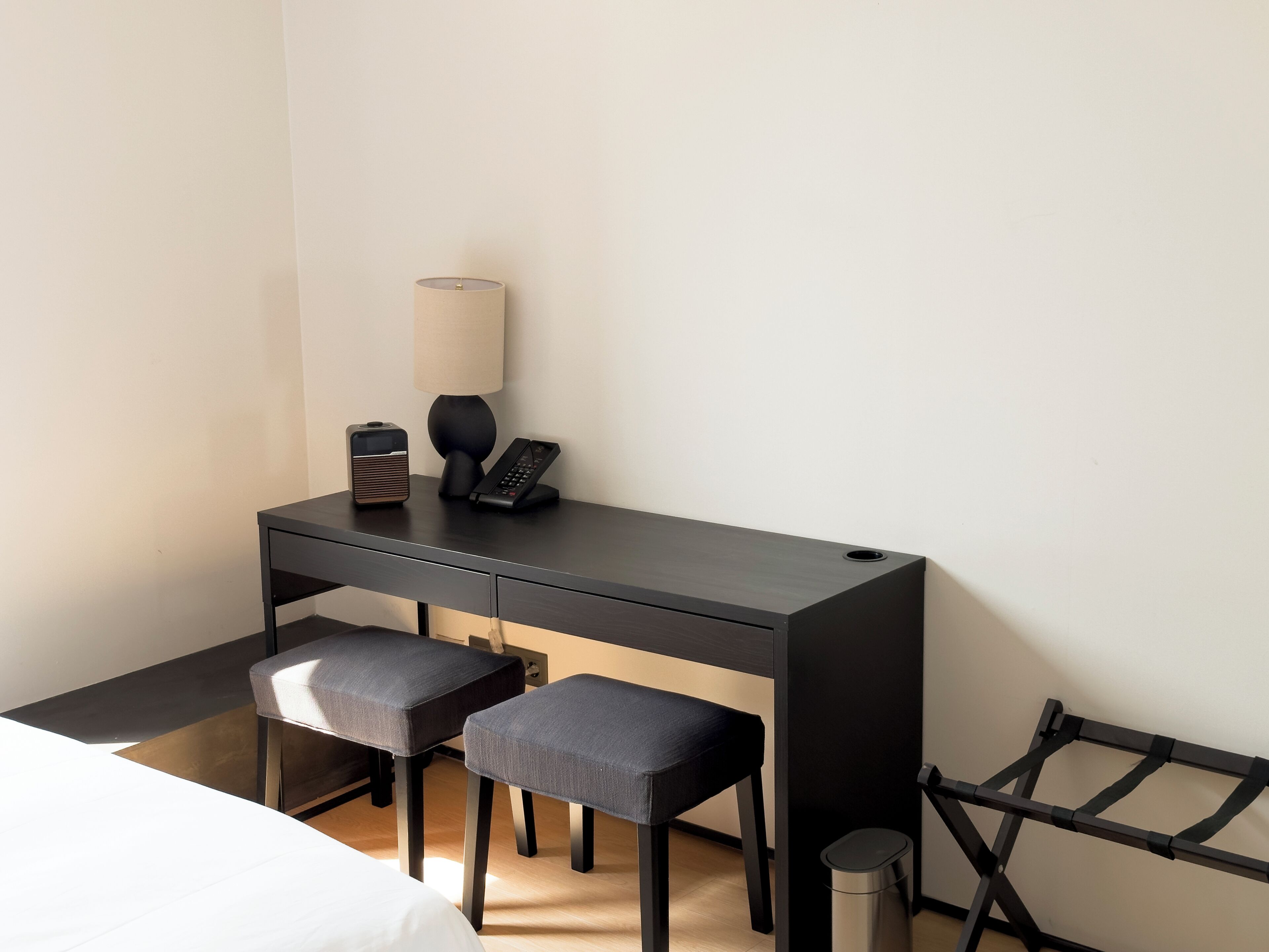 A compact hotel room with a dark wooden desk, two matching chairs, a bed, a lamp, a telephone, and a trash bin.
