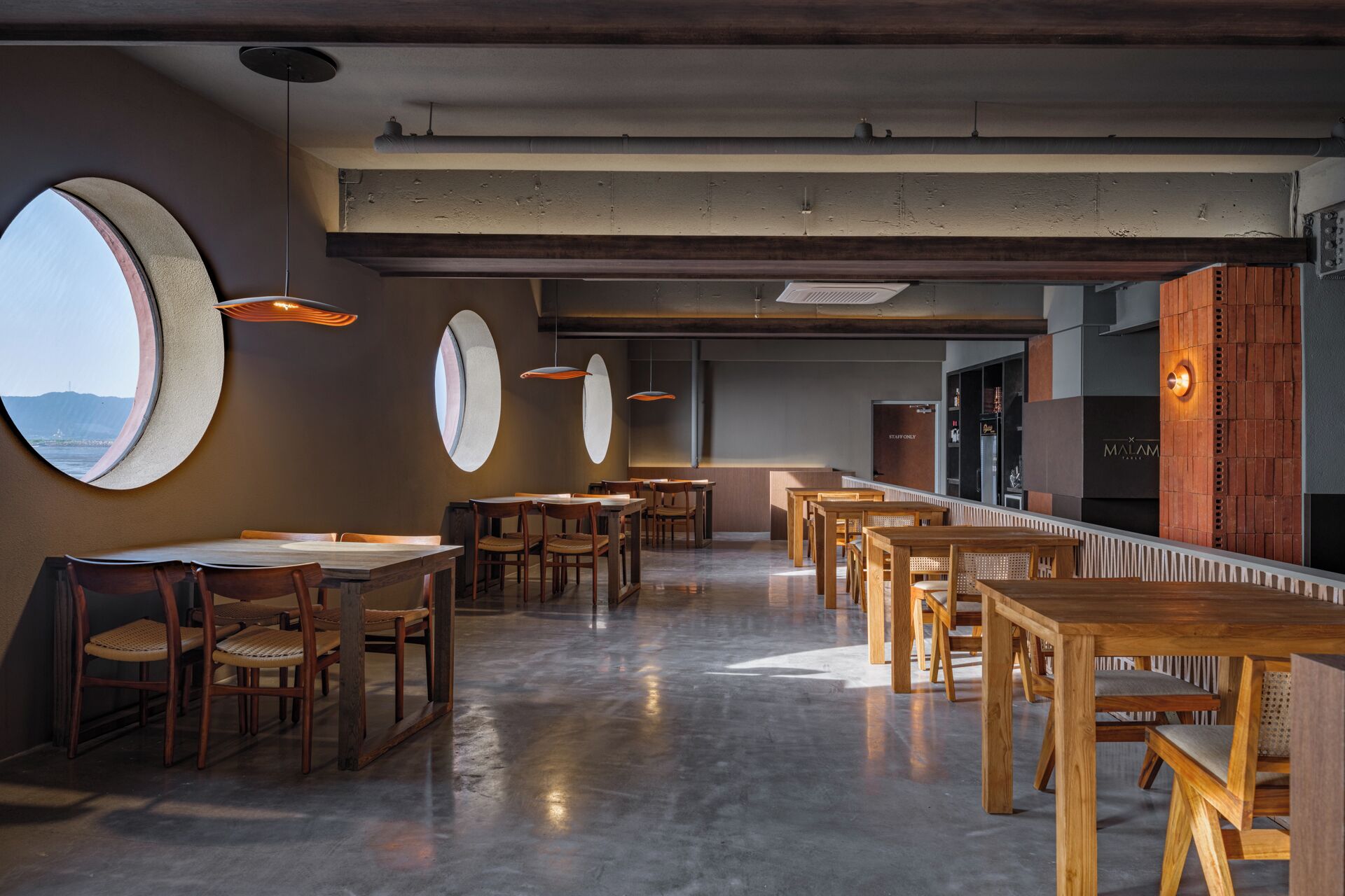 A modern restaurant interior with round tables and chairs, a concrete floor, and large windows offering views of the sea.