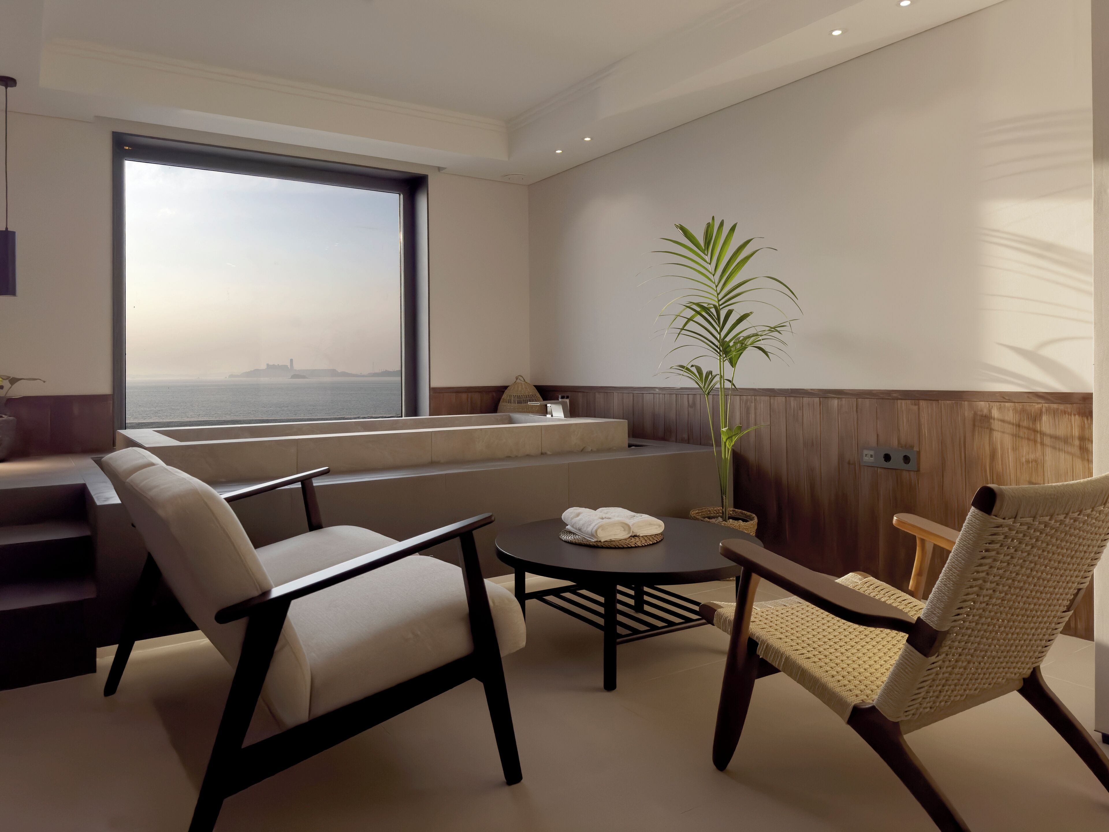 A modern, well-lit room with a large window overlooking a body of water, a seating area with a table, and a potted plant.