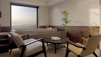 A modern, well-lit room with a large window overlooking a body of water, a seating area with a table, and a potted plant.
