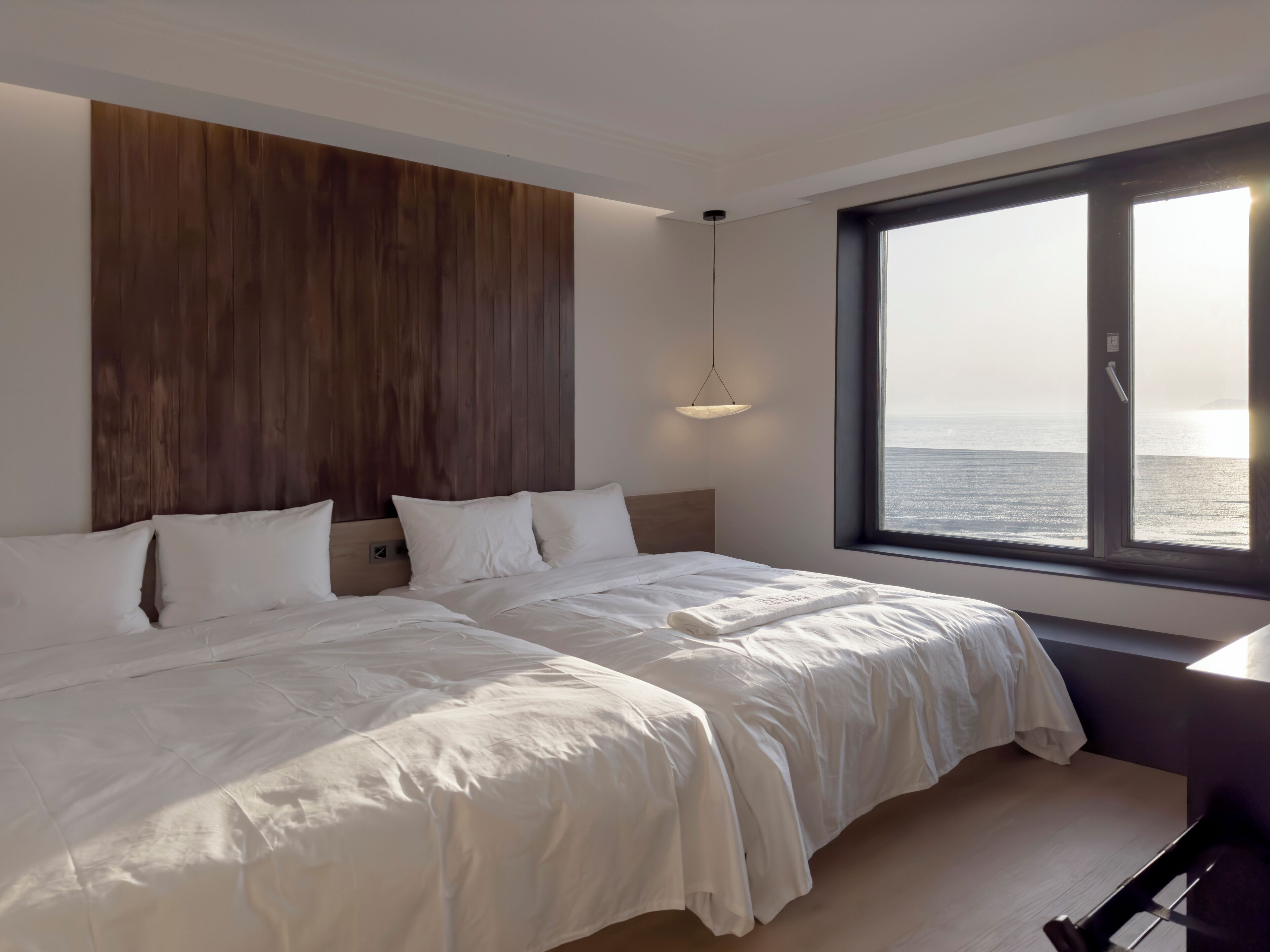 A modern hotel room with a large bed, a wooden headboard, a bedside table, and a view of the sea through a large window.