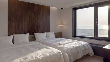 A modern hotel room with a large bed, a wooden headboard, a bedside table, and a view of the sea through a large window.
