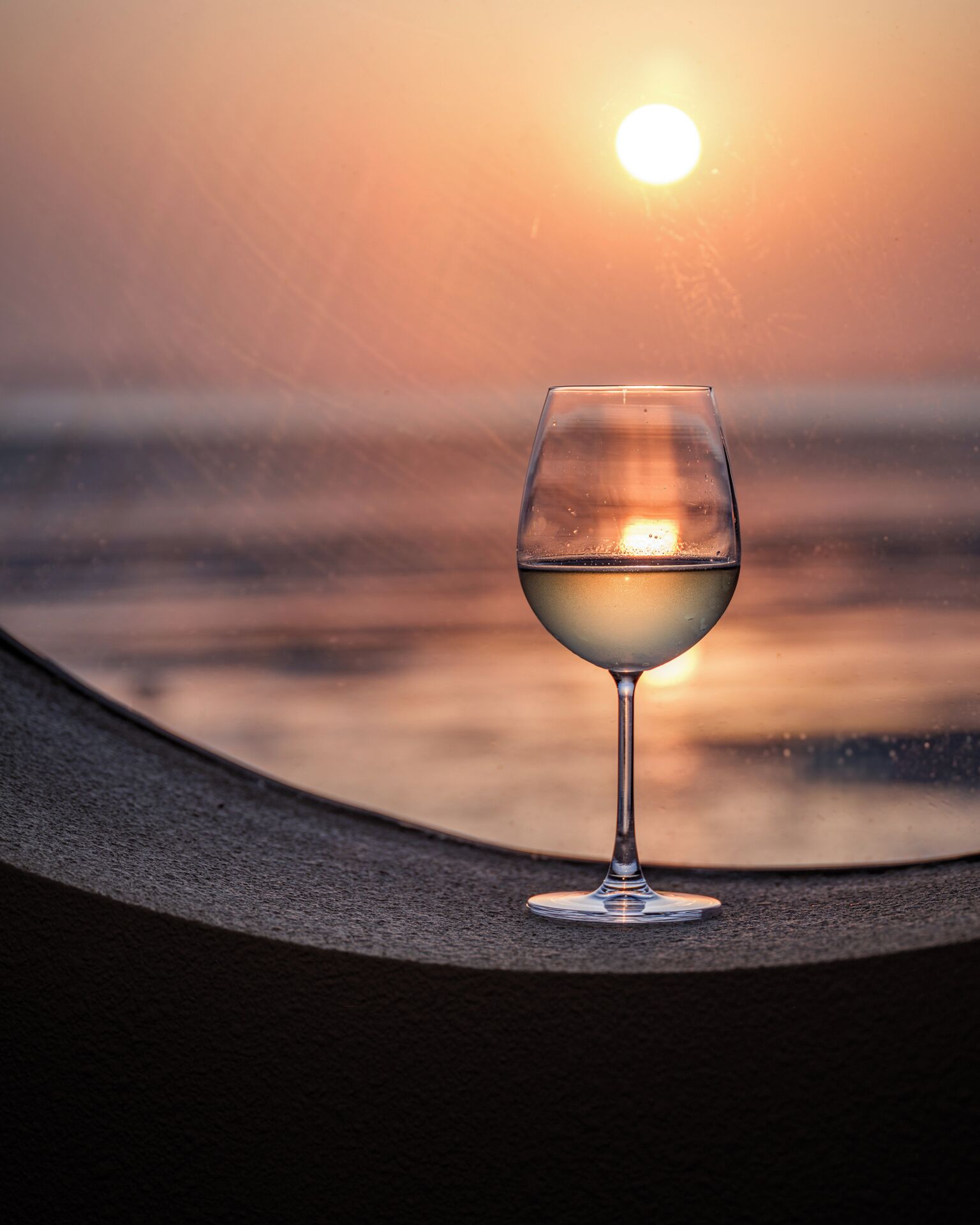 A glass of white wine on a curved ledge with a sunset in the background.