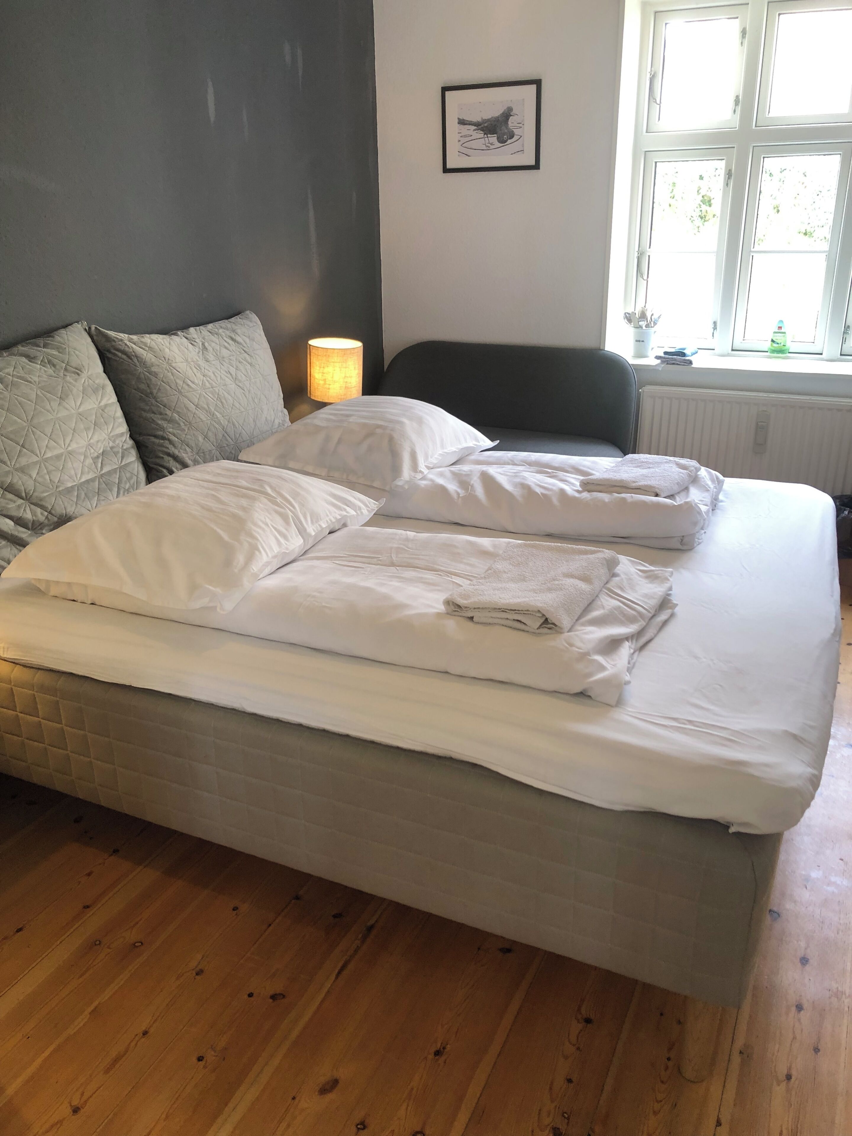 Ein Doppelbett mit weißen Leinen, eine Nachttischlampe, ein gerahmtes Bild an der Wand und ein Fenster mit Blick ins Grüne.