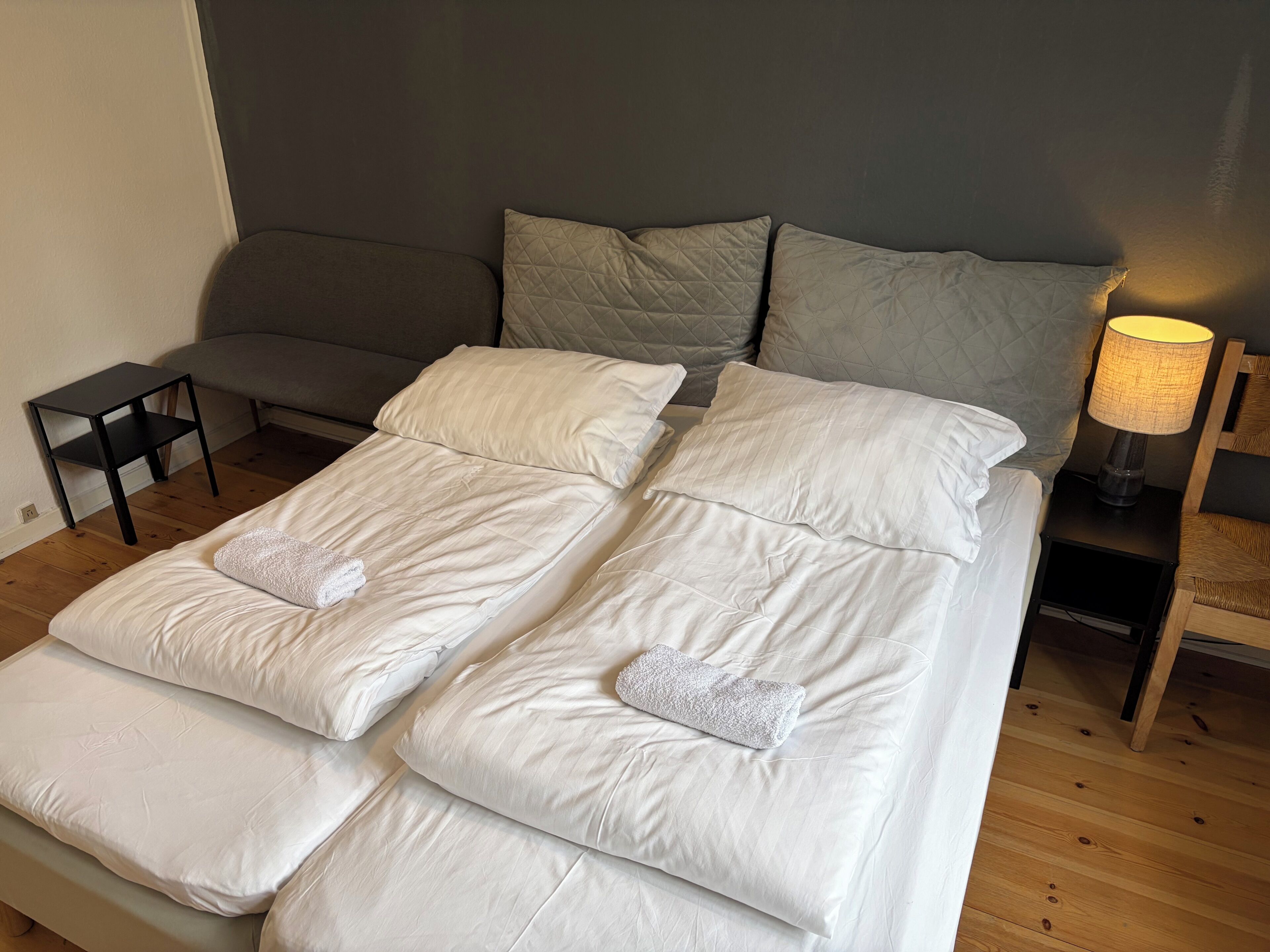Una cama doble con ropa de cama blanca, una mesita de noche, una lámpara y un sofá gris en una habitación con suelo de madera.