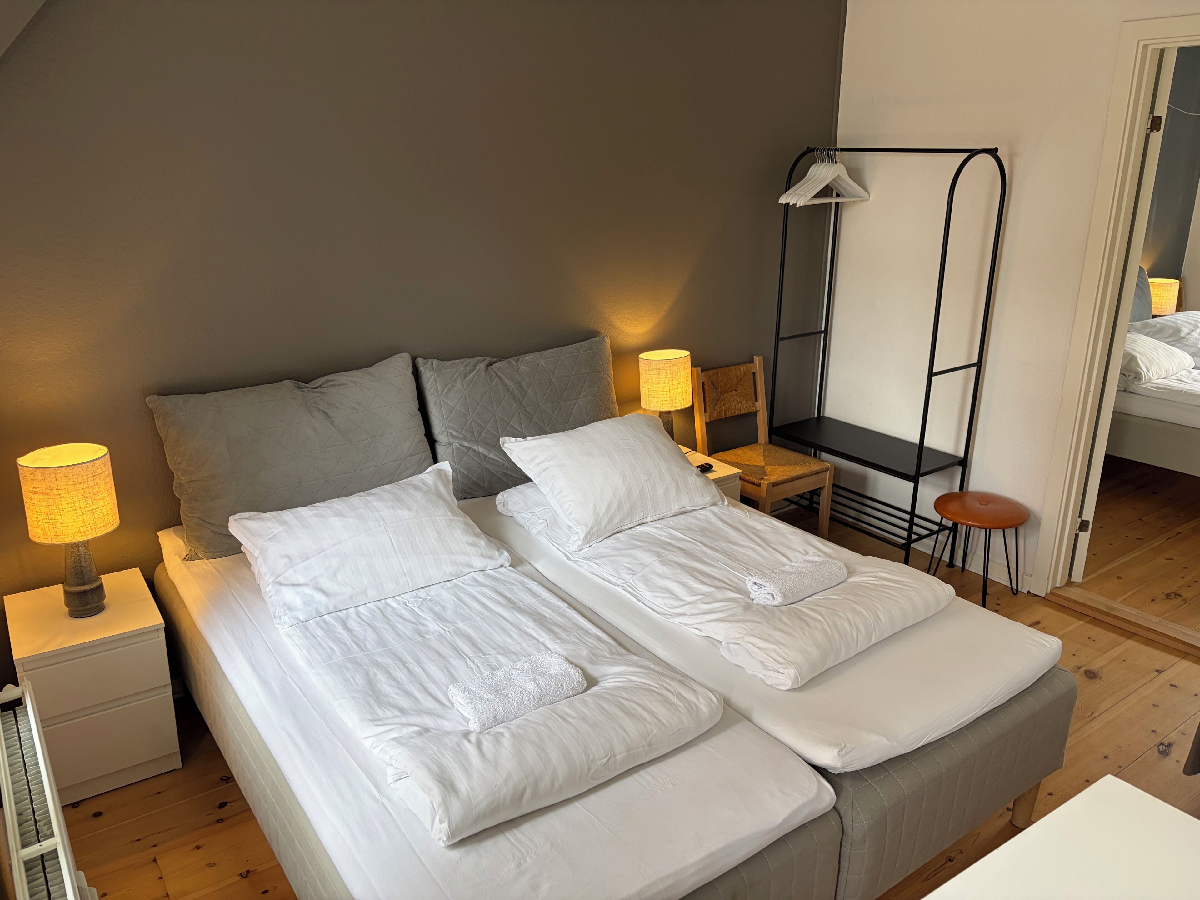 Un lit double avec du linge de lit blanc, deux tables de chevet avec des lampes, une chaise en bois et un petit tabouret.