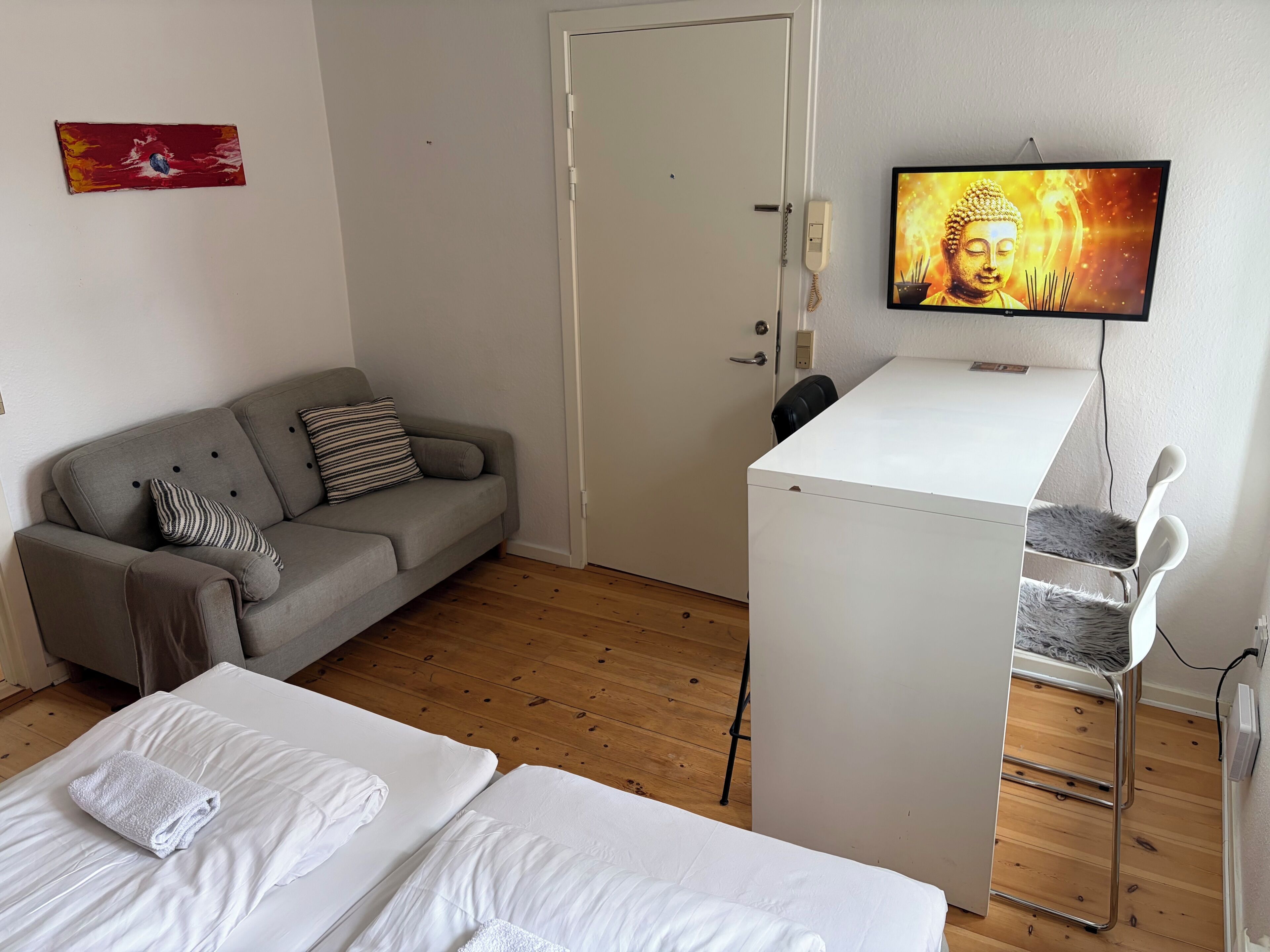 A room with a grey sofa, a white desk, a television, and a bed with white linens.