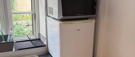 Una kitchenette compacta con microondas, refrigerador y cafetera sobre una repisa de metal negro.