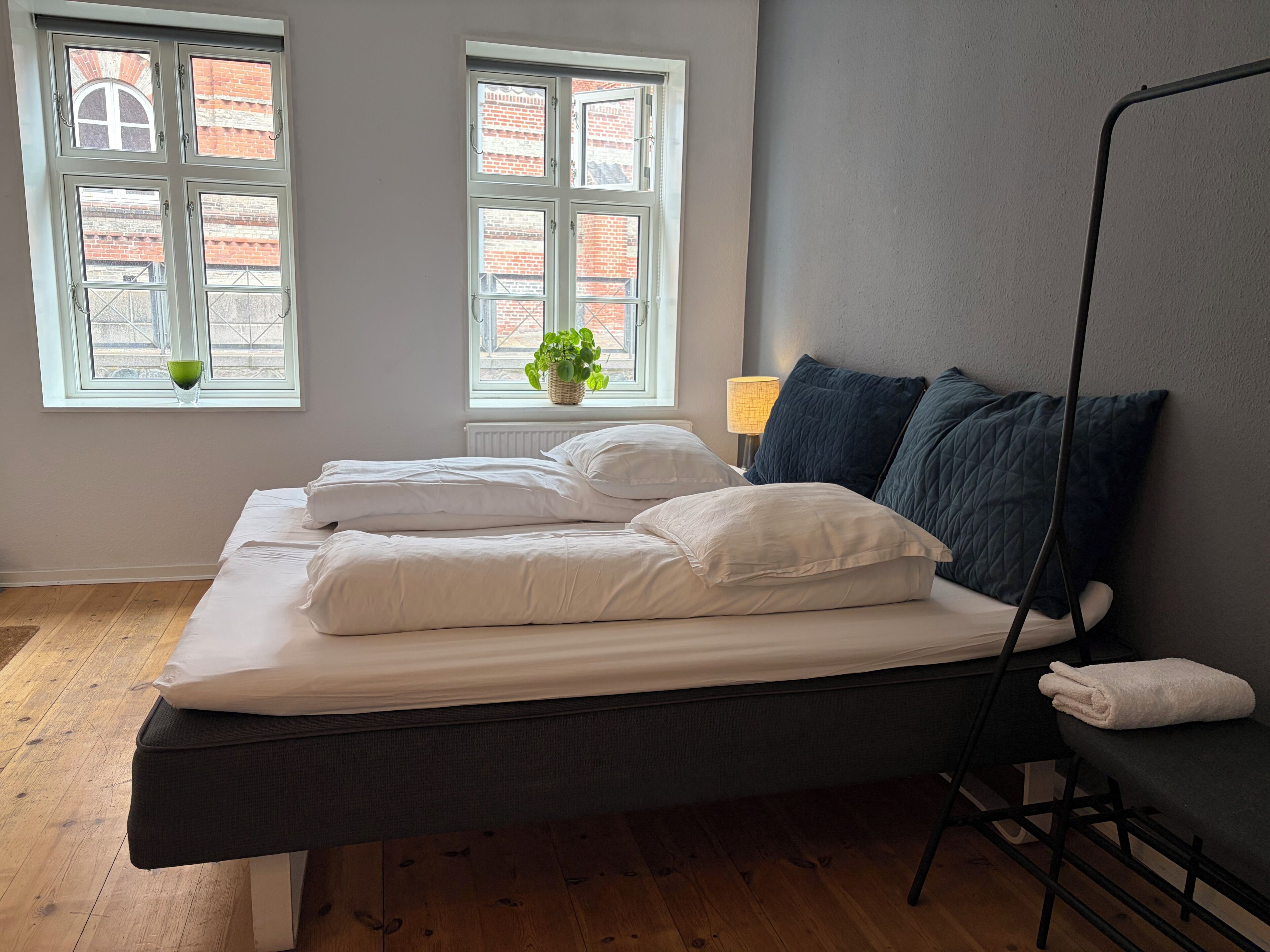 Quarto com uma cama, mesa de cabeceira, abajur e duas janelas.