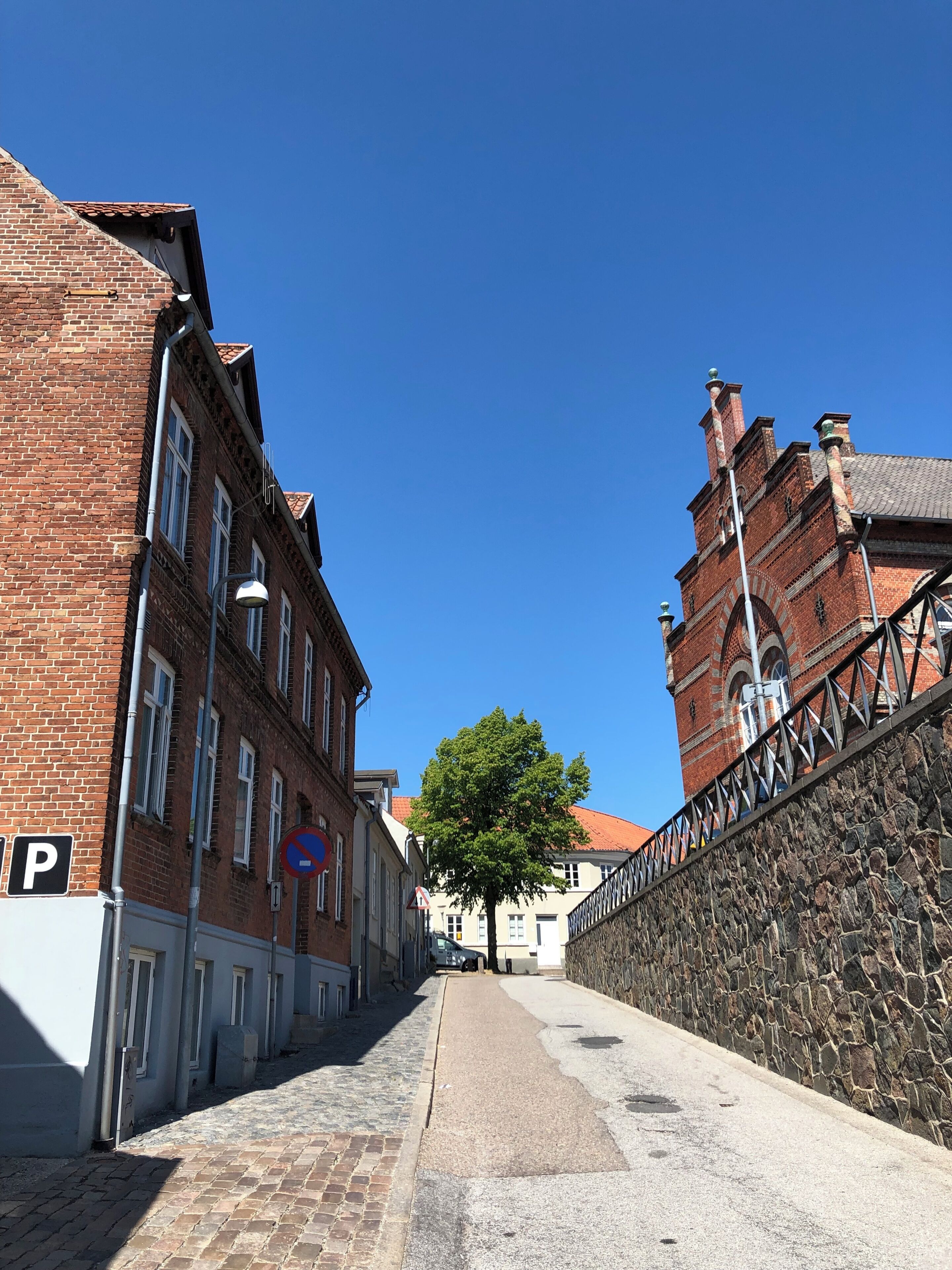 A narrow street with brick buildings, a stone wall, and a clear blue sky.