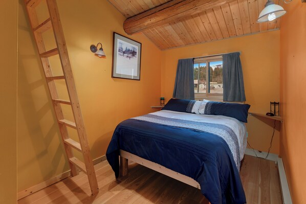 A bedroom with a wooden ladder, a bed with blue bedding, a framed picture on the wall, and a window with curtains.